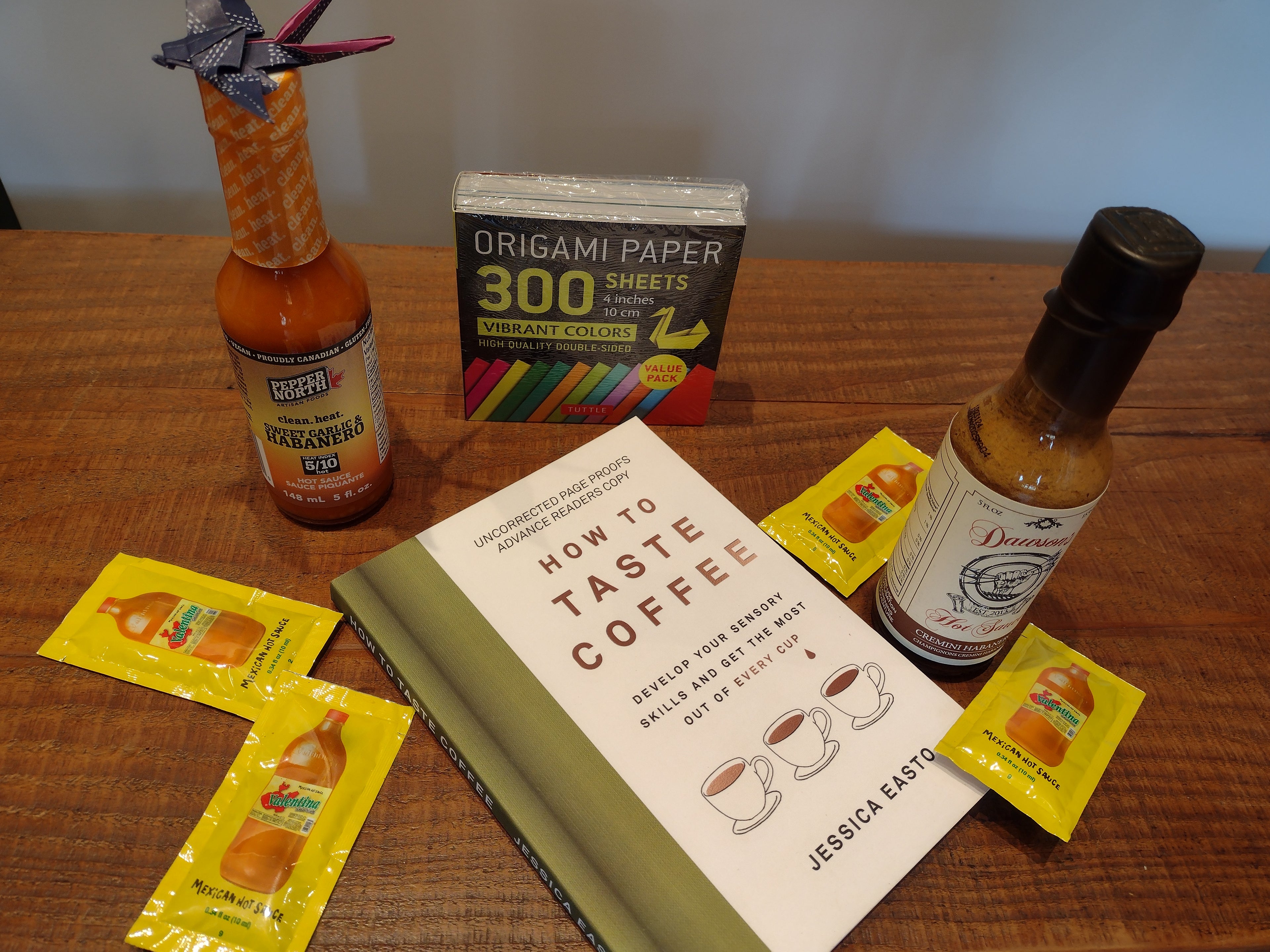 the book How to Taste Coffee, a package of 300ct 10cm Tuttle Origami paper, a bottle of Pepper North Habanero hot sauce, a bottle of Dawsons's Cremini hot sauce and packets of Valentina's hot sauce scattered across a wooden surface.