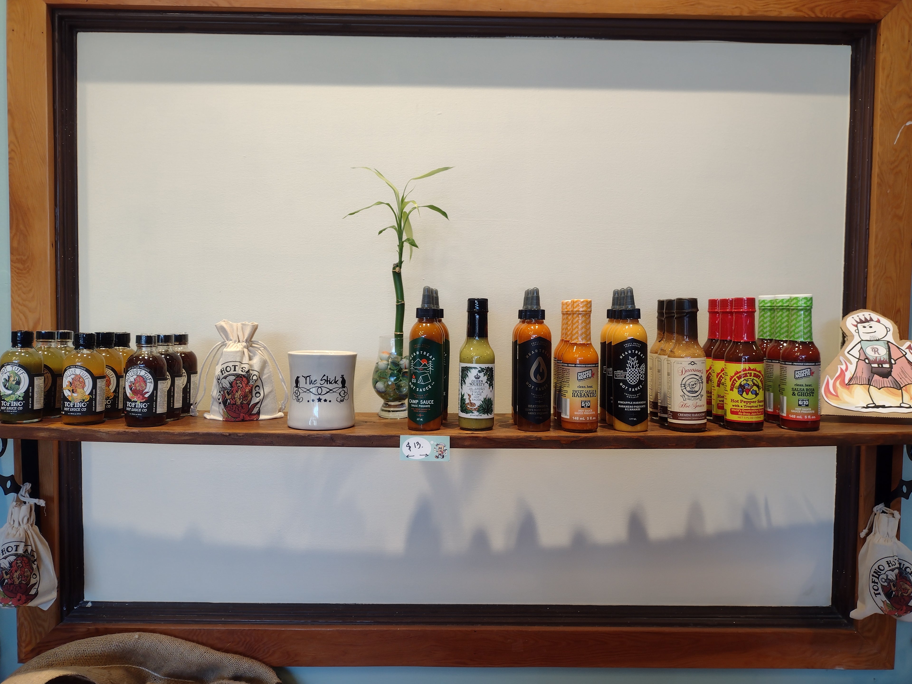 A thin wooden shelf full of hot sauce bottles of different brands with a bamboo plant and a cardboard cutout of a cartoon stick figure wearing a cape standing in front of flames flames.