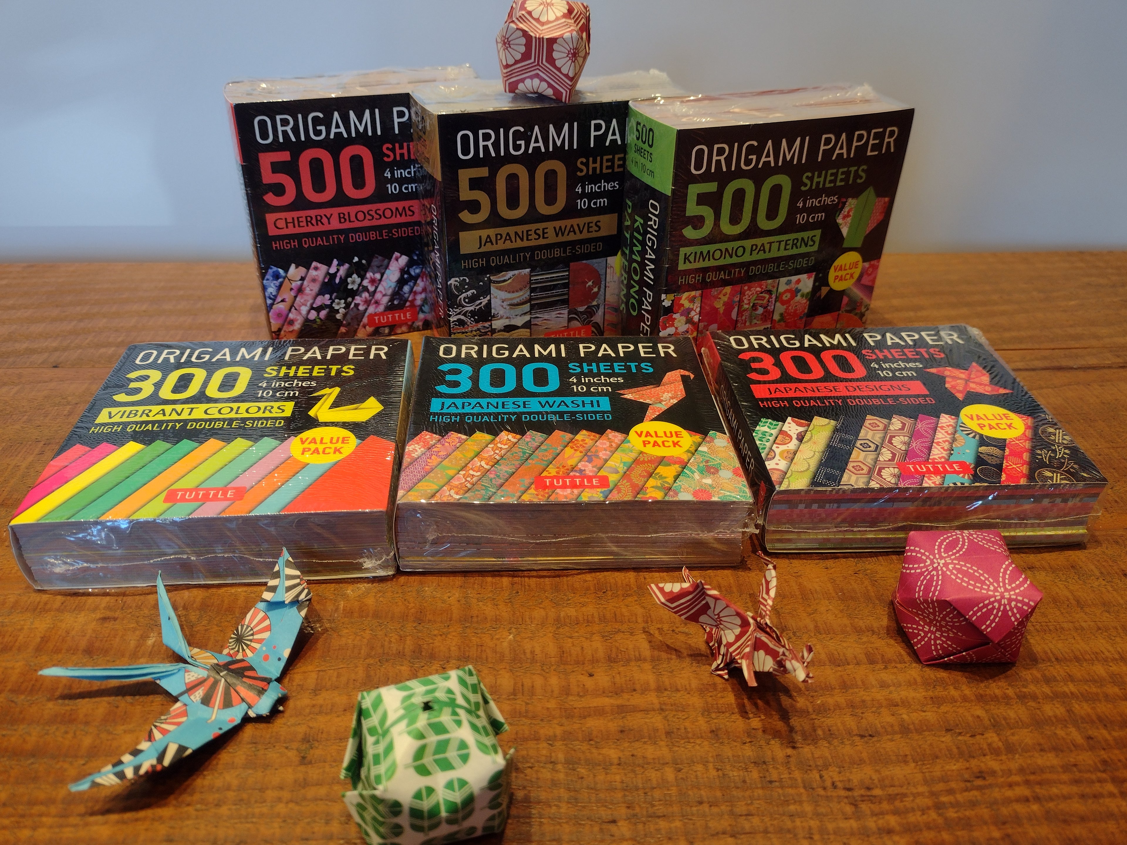300ct and 500ct wrapped packages of Tuttle origami paper on a wooden surface with constructed origami models scattered about